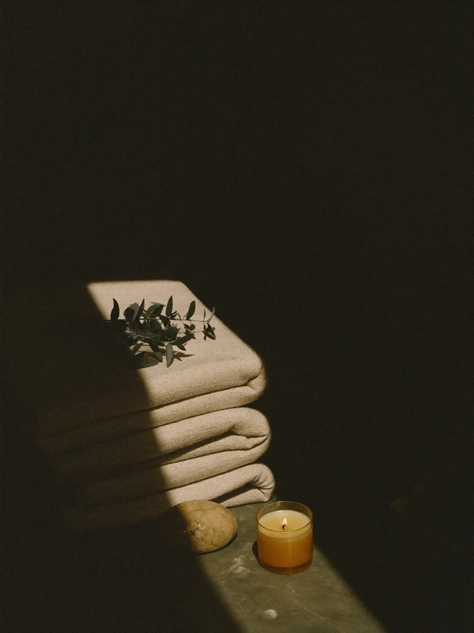Folded natural linen with a sprig of eucalyptus, a smooth stone and a lit candle in a single beam of warm light.
