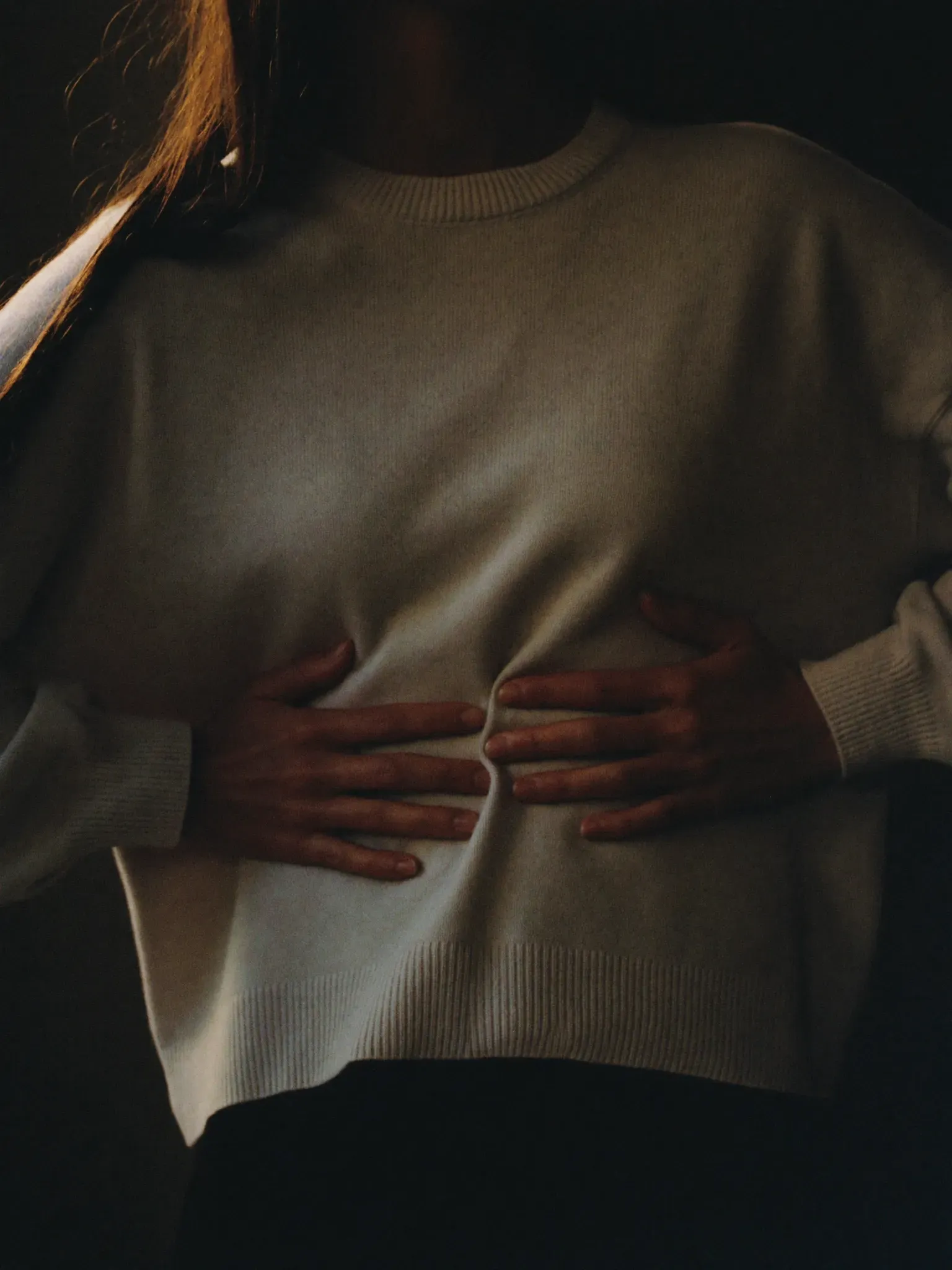 Hands resting on the lower ribs over a cream knit sweater, mid slow breath, soft window light.