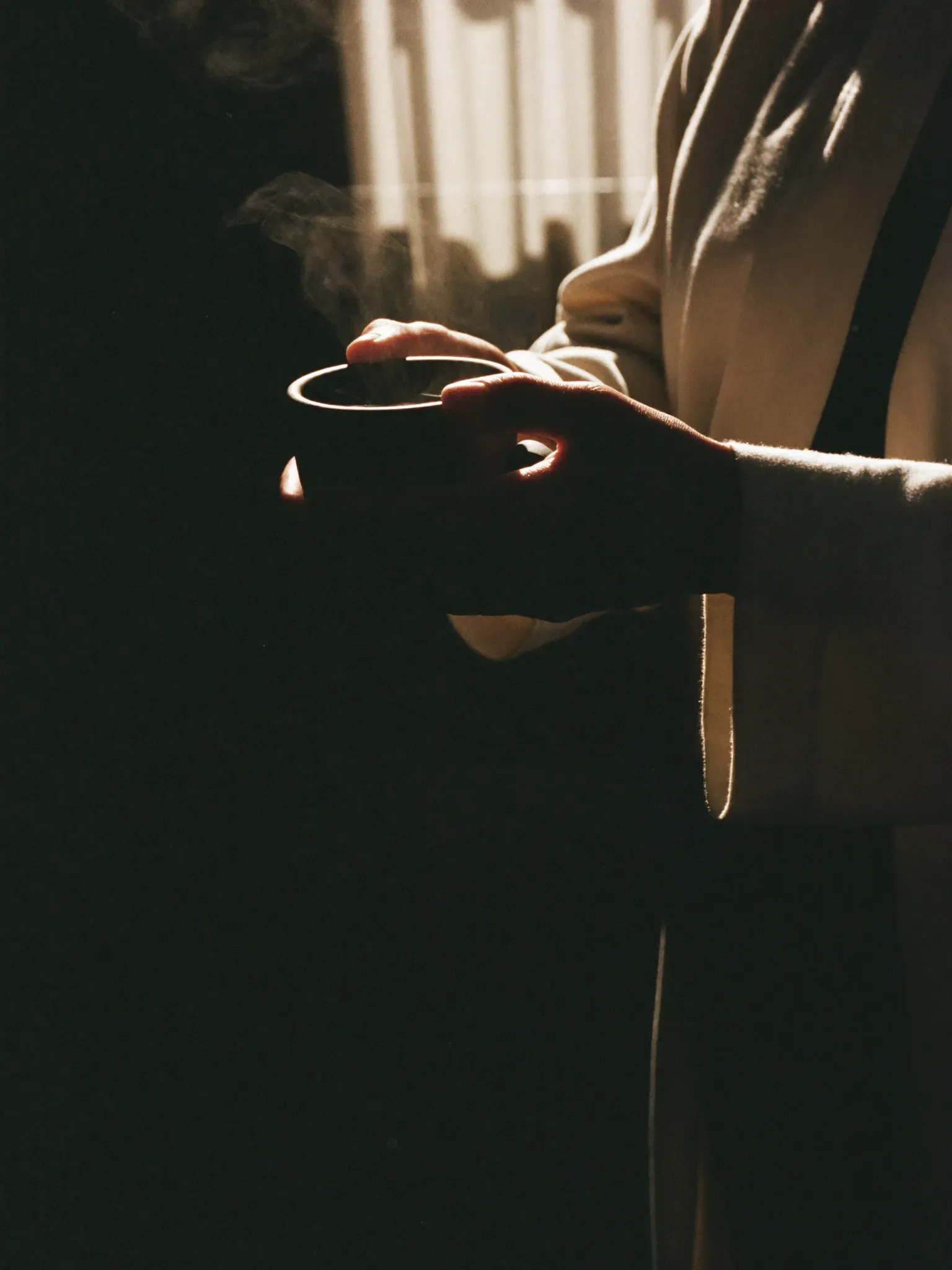 Hands wrapped around a steaming ceramic cup, a cream linen robe sleeve, early morning sun through sheer curtains.