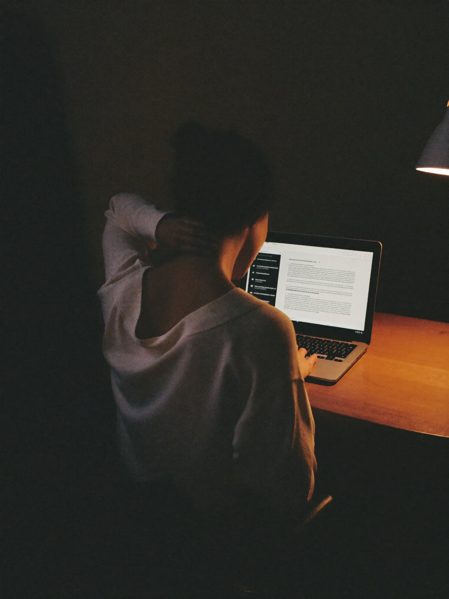 A woman at a laptop late in the evening, rubbing the back of her stiff neck, shoulders rounded forward.