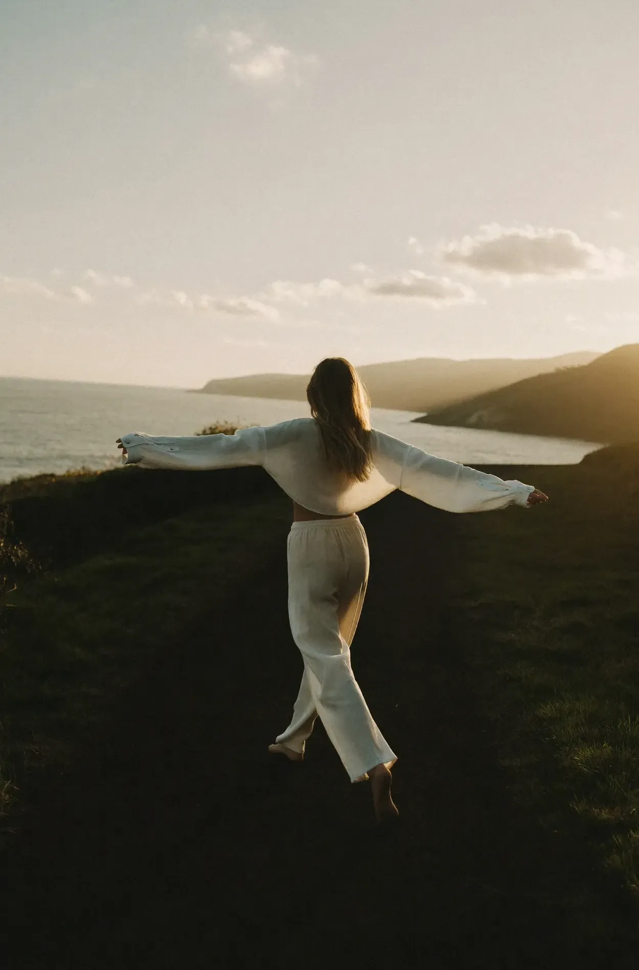 A woman in loose cream linen walking barefoot along a coastal path at golden hour, arms open wide.